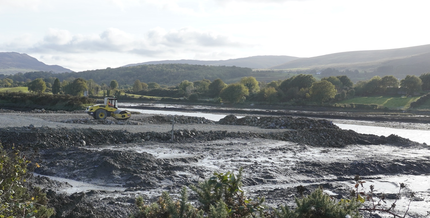 View over water with shore on left. Ahead
                          is a road roller on some rocks that have been
                          pushed out onto the mud flats.