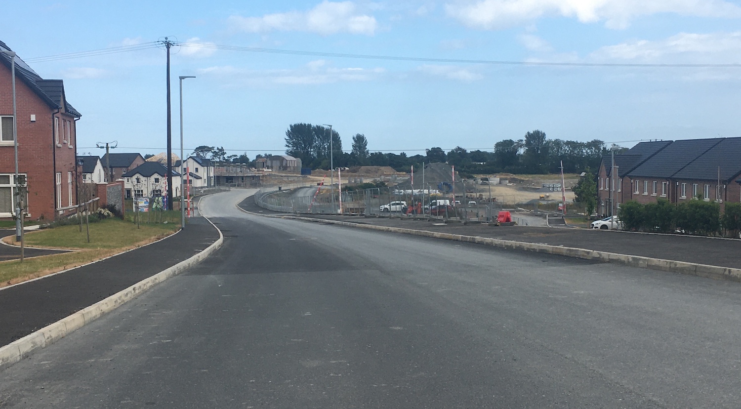 View along a road that has been freshly
                          tarmacked, with a completed house on the left
                          and incomplete houses ahead.