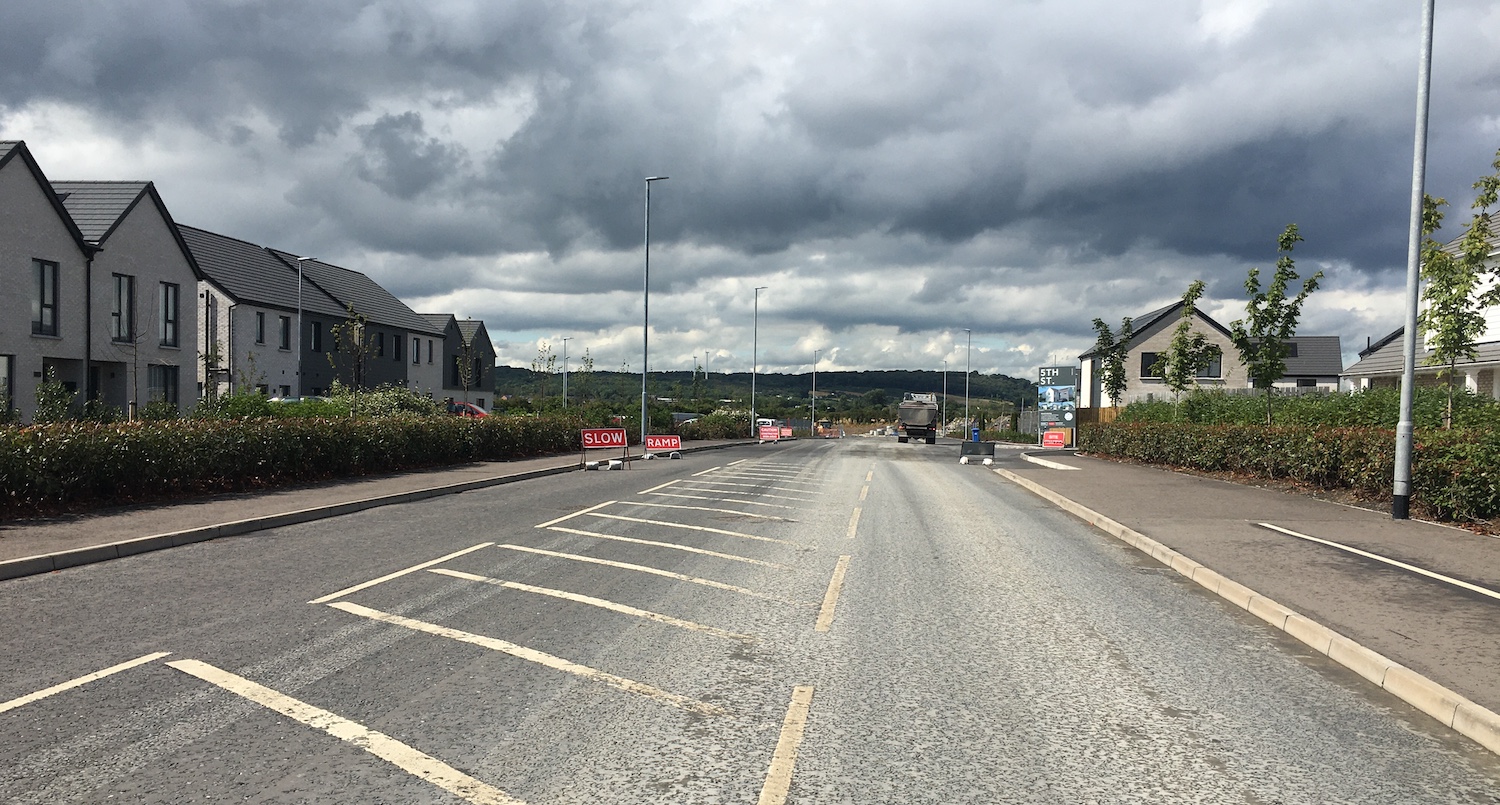 View along a completed road with harching
                          in the centre, but with evidence of house
                          construction up ahead in the form of plant
                          equipment.
