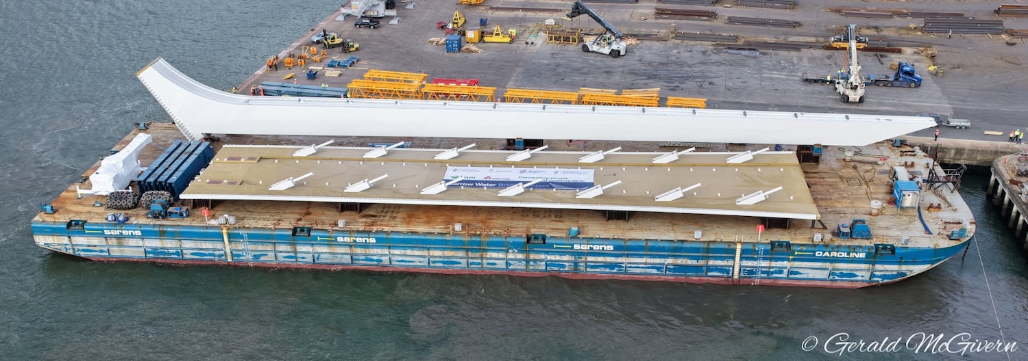 Aerial view of a barge. Two items are
                          side by side. The further one is a long
                          needle-like metal structure, thinner at the
                          right than the left. The nearer one is a piece
                          of road deck with 7 cable connectors on each
                          side.